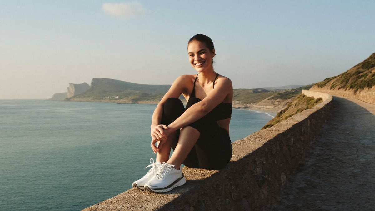 A woman resting on a low wall above the sea after a run, smiling out at the water