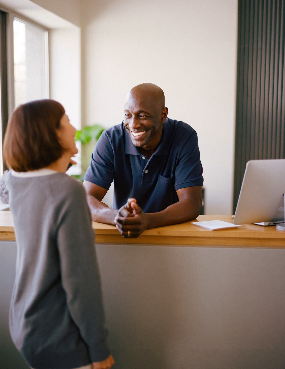A chiropractor leans on the reception desk of a bright clinic, smiling as he greets a patient.